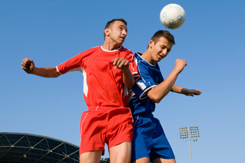 Two football players, one is dressed in a red top and shorts, the other in a blue top and shorts. The one in blue is heading a white football.