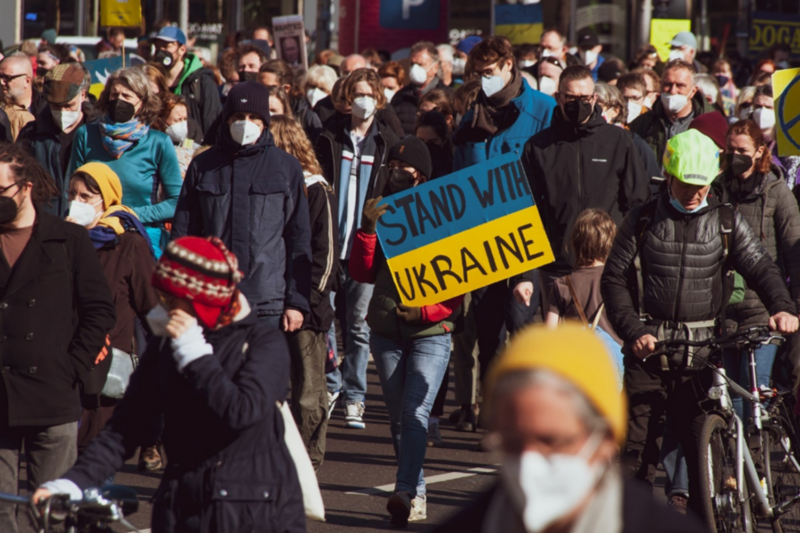 Crowd of people marching in protest, one person holds a poster with the Ukrainian flag which says 'Stand with Ukraine'
