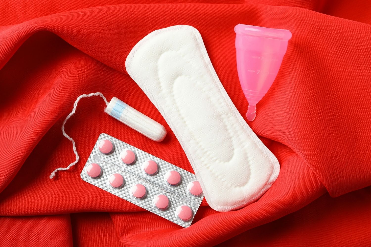 Image of a contraceptive pills, a tampon, pad, and menstrual cup on a red material background.