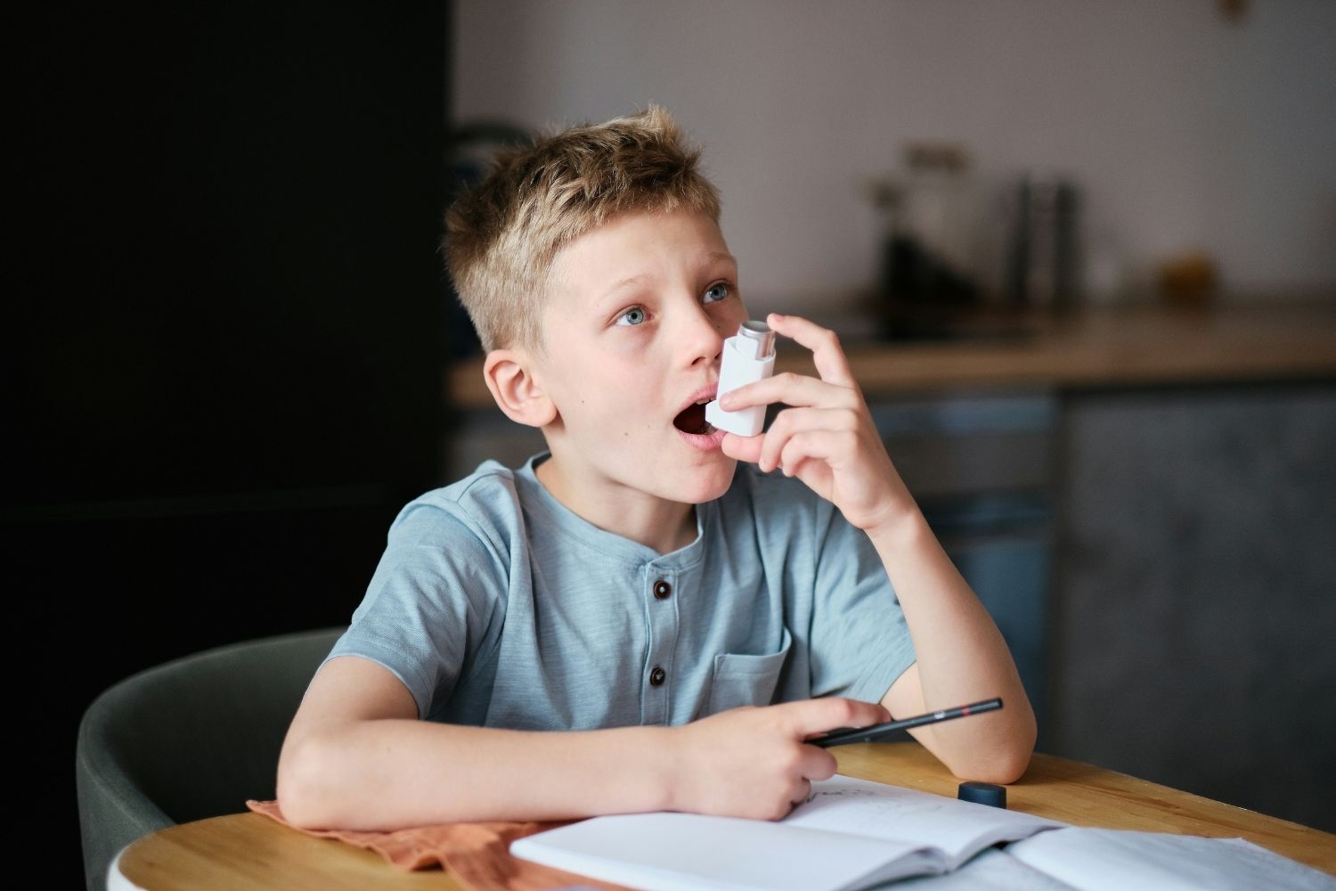 Young boy uses an asthma inhaler whilst sat at a table.