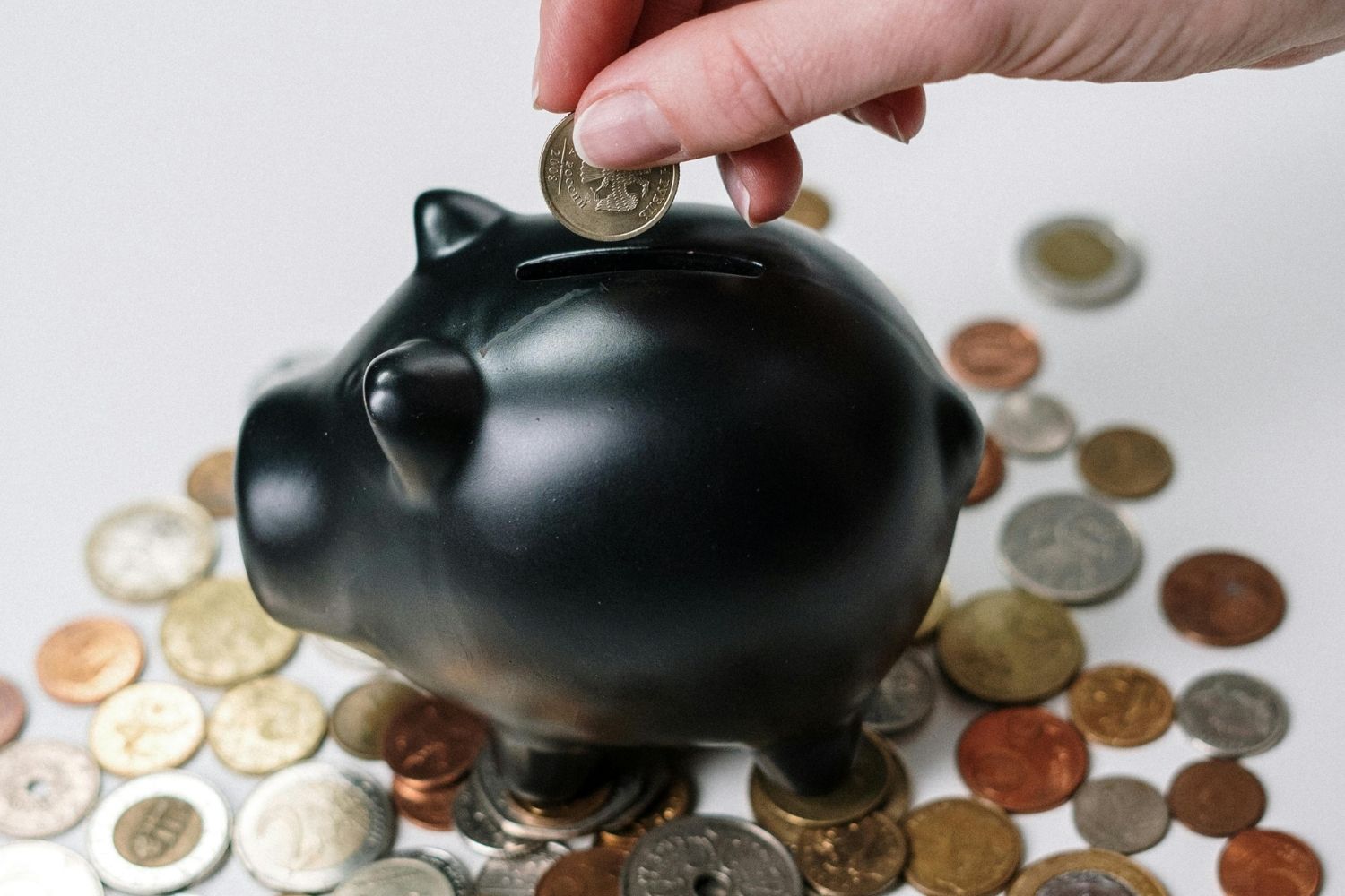 Person putting a coin into a blank piggy bank. The piggy bank sits on top of a pile of coins.