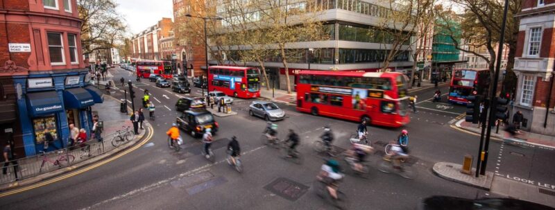 Photograph of a road junction in London with 4 red buses, a few taxis and lots of cyclists.