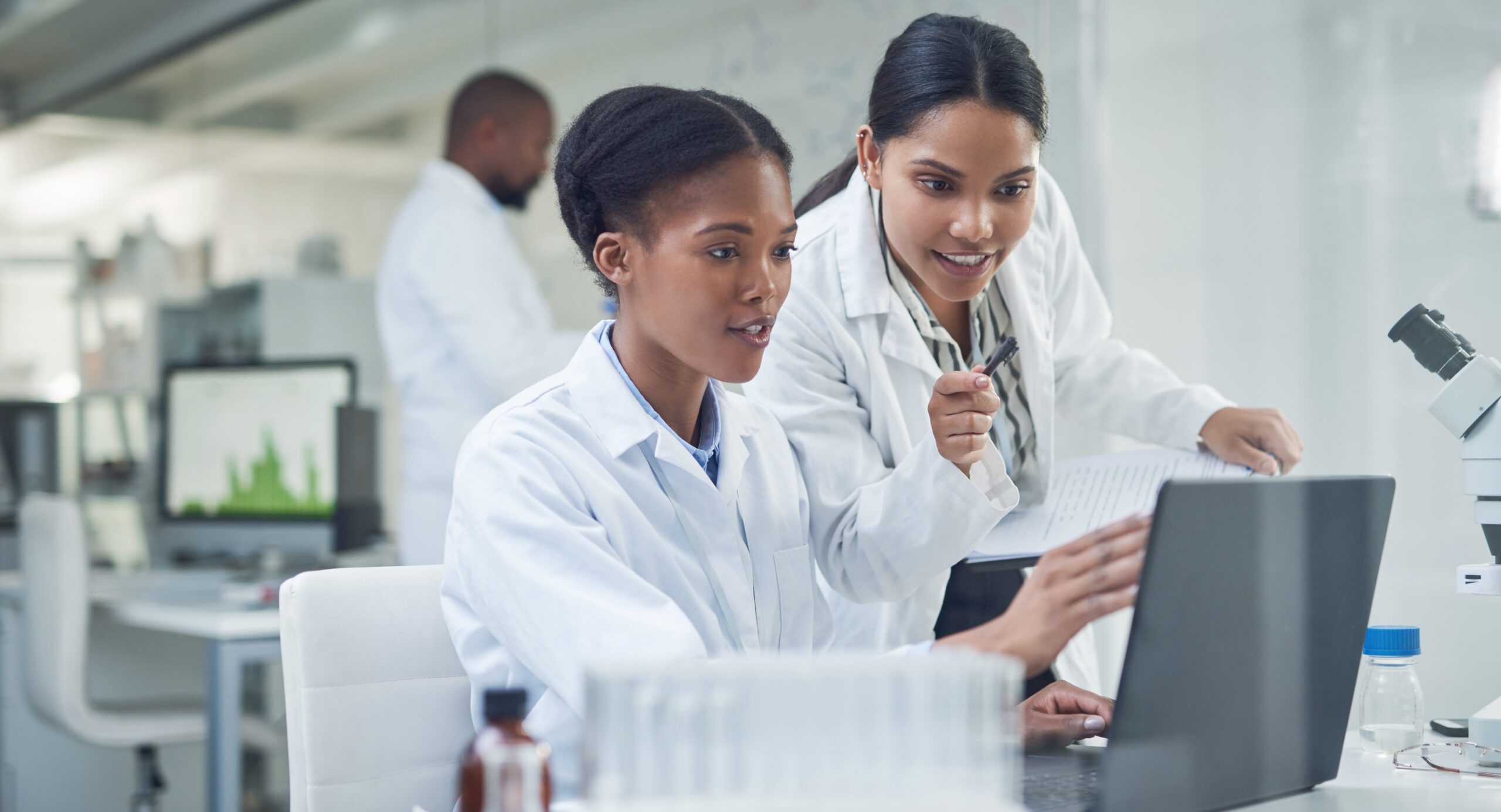 Two researchers in a lab, wearing white coats, look at a laptop screen.