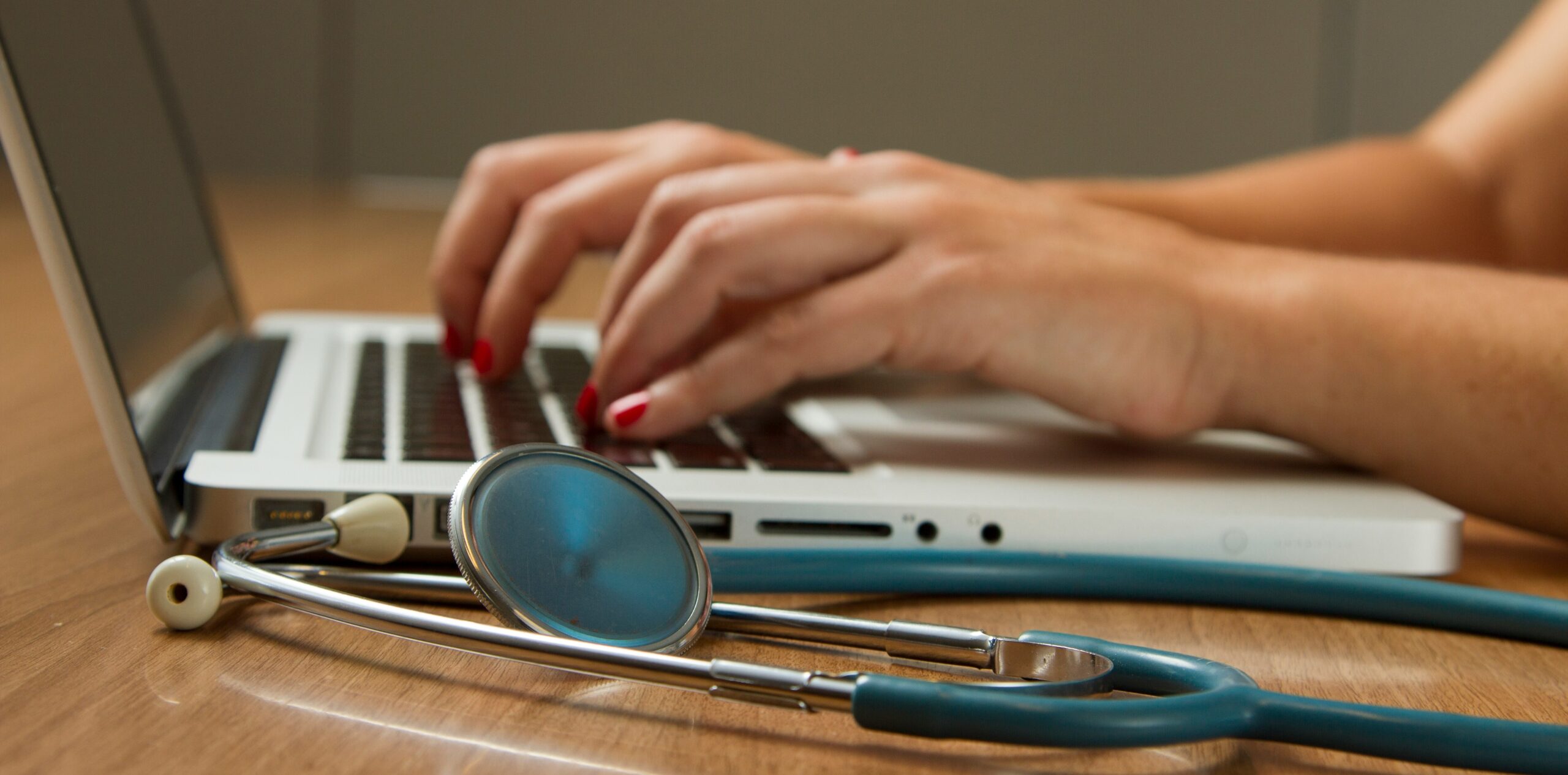 Photo of someone's hands at they type on a laptop. A stethoscope is next to the laptop.