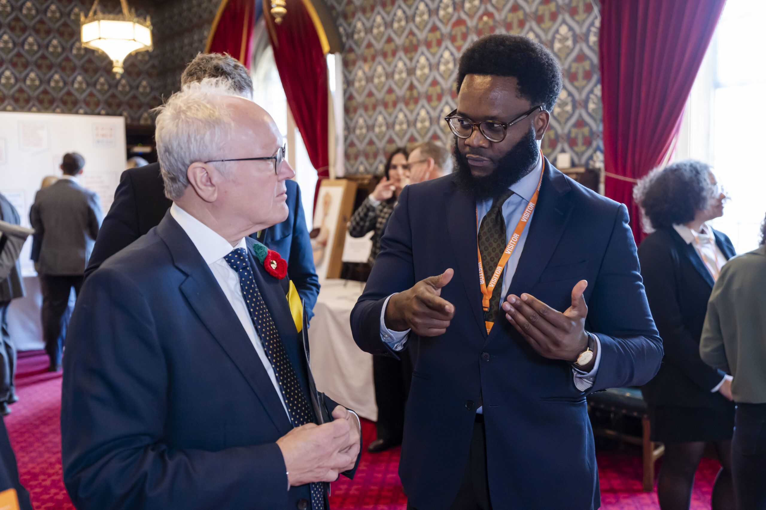 Two people wearing suits in discussion. They are standing in a busy room within the Palace of Westminster.