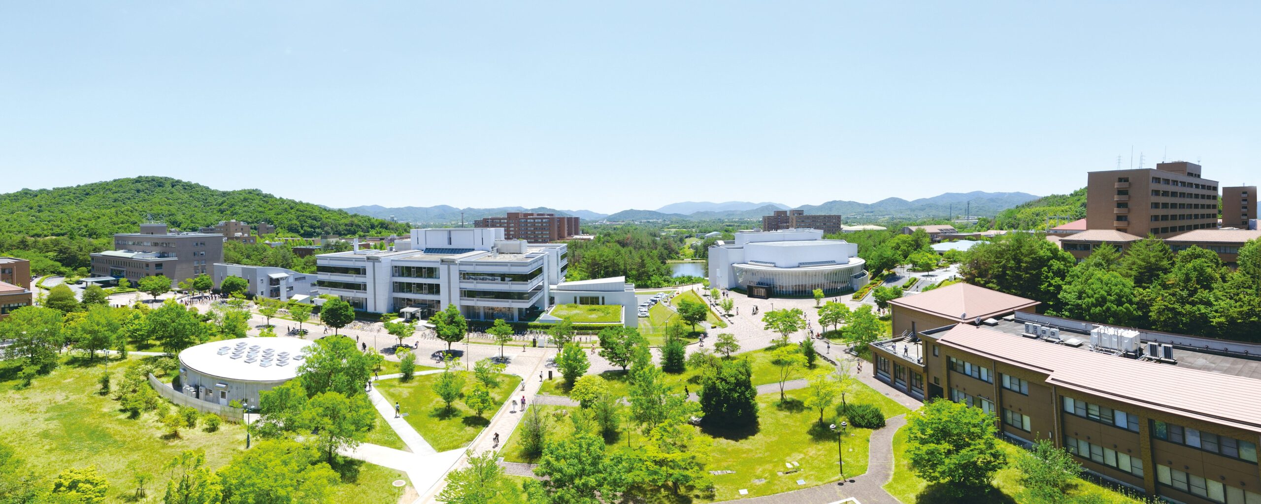 A panoramic photo of the Hiroshima University campus. Clear blue sky, paths through lawns and trees, and buildings of various styles.