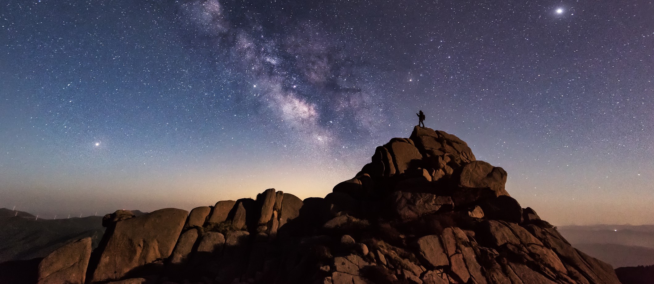 Silhouette of a person standing on top of a rocky mountain with a star-filled night sky behind them.