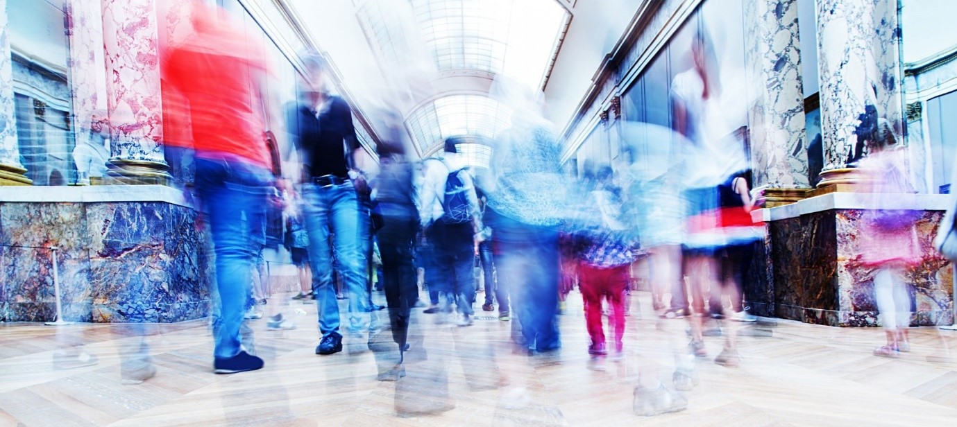 Blurred photograph capturing high foot traffic in an indoor public space, with marble columns, glass roof and a wooden floor.
