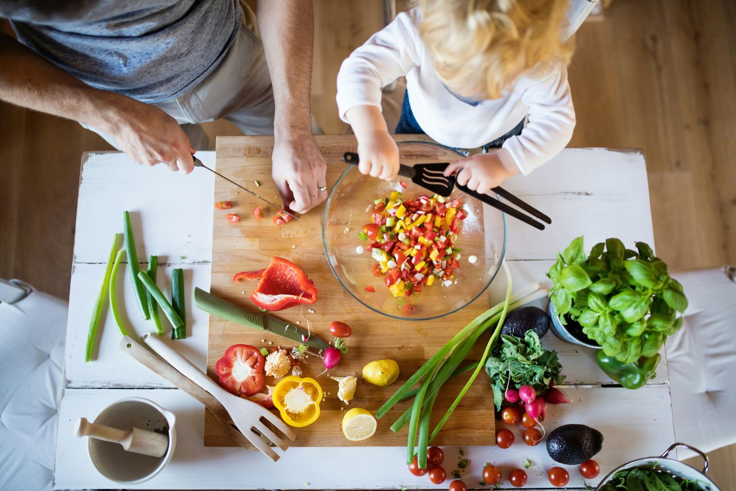 A top down photo of an adult and a child cooking with some vegetables. A bell pepper has been cut up and put into a glass bowl which is being mixed by the child.