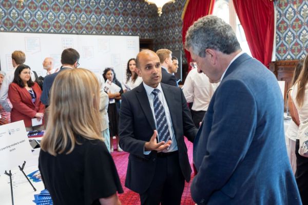 Three standing people talking in a large busy room. In the background the walls have highly-decorated wallpaper and there's a grand window with red curtains.
