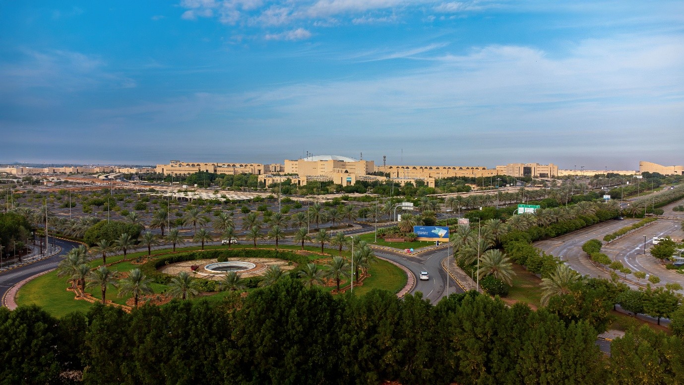 Landscape photo of Qassim University, with trees and a large roundabout in the foreground and yellow-stone buildings behind.
