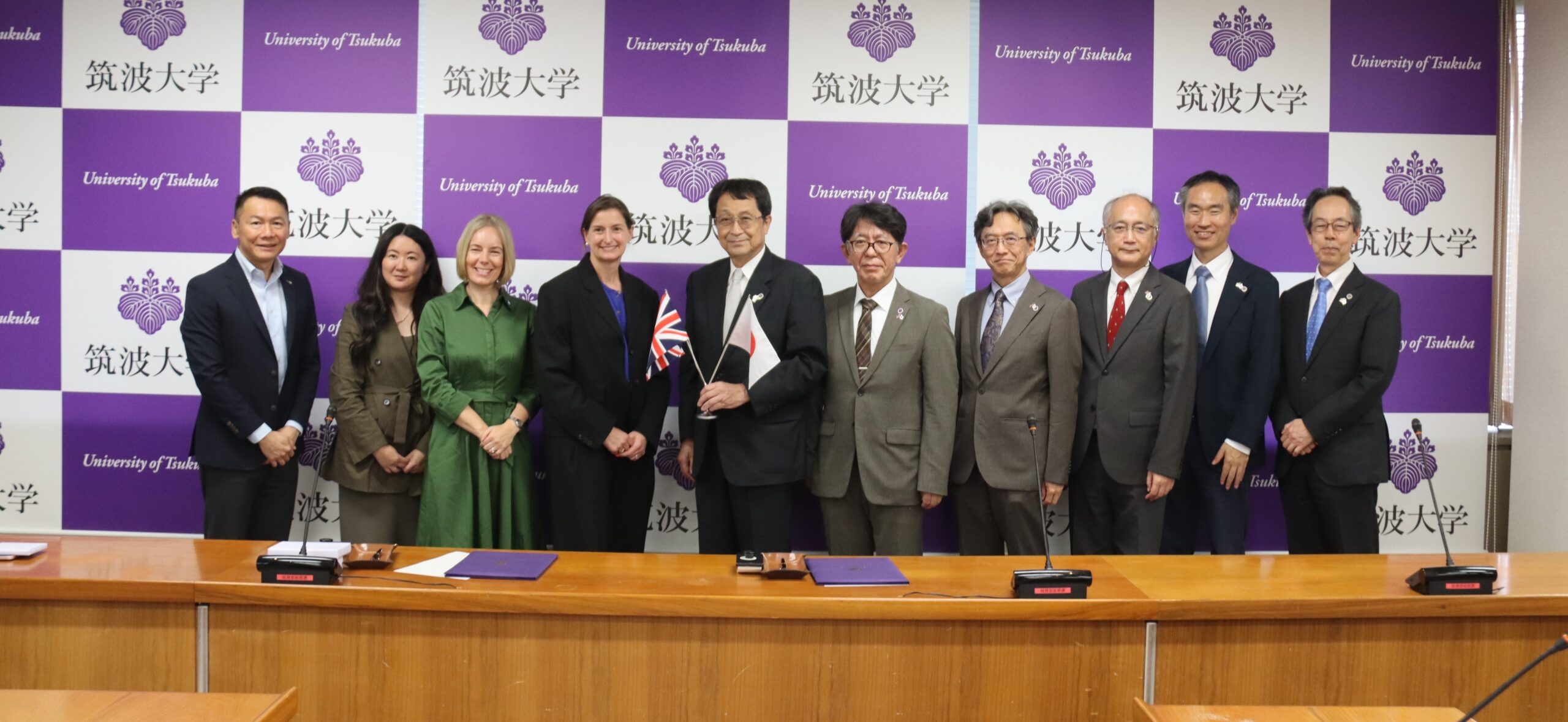 10 smartly-dressed people standing in a row behind a long desk. The person in the middle is holding the Japanese and British flags. Behind them is a screen with the University of Tsukuba logo repeated.